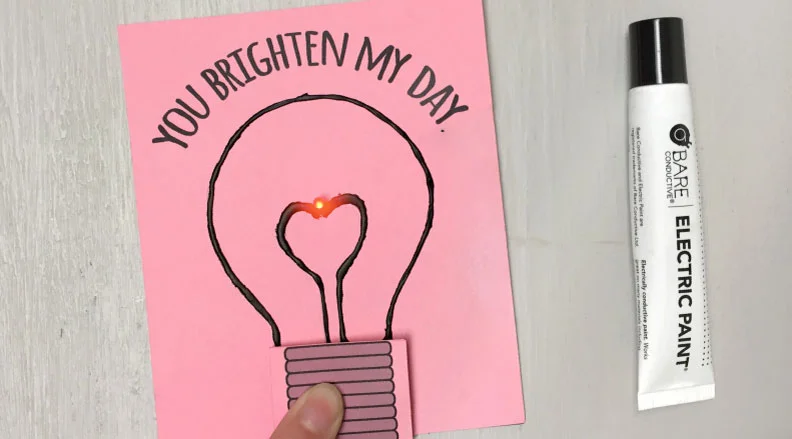 Hand holding a card that reads 'You Brighten My Day' with a light-up heart. A black electric paint pen is placed next to it