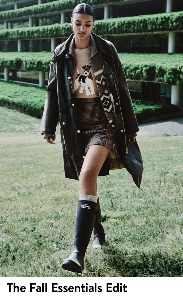 A woman wearing a waxed jacket and knit cardigan over a sweater with an image of a dog, a miniskirt and rain boots.