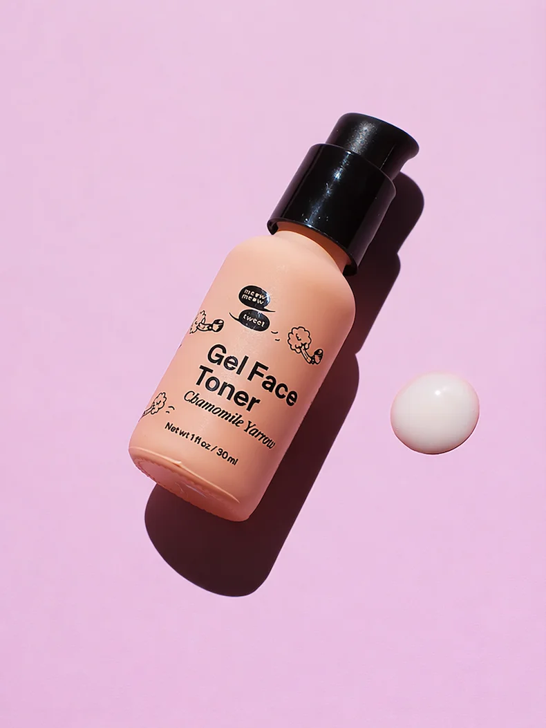 A pink bottle of gel toner is lying down on a pink backdrop with a drop of white gel toner next to it.