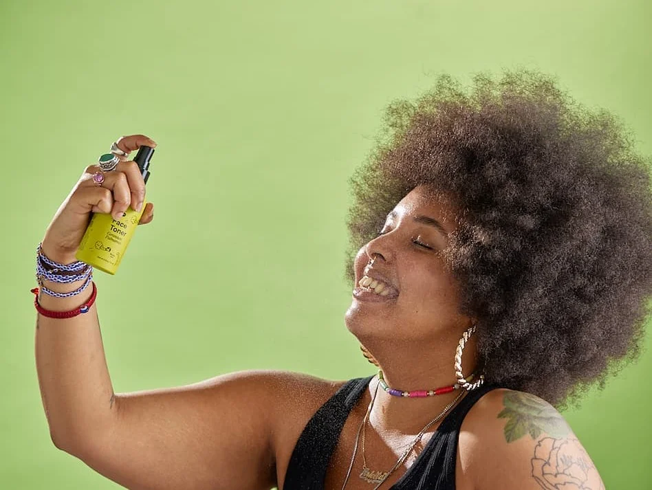 A person against a green backdrop mists their face with green spray bottle.