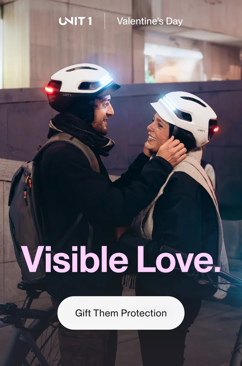 Couple wearing UNIT 1 smart helmets with front and rear lights illuminated, standing with their bikes and smiling at each other in an urban setting at night.
