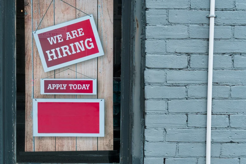 A window in a grey brick building with “We are hiring” and “Apply today” signs hanging in it.