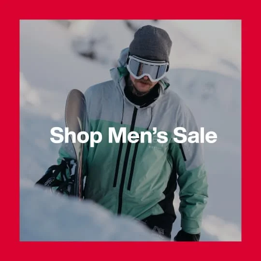 A darkened image of a Burton walking in snow carrying a Burton Snowboard wearing the [ak] Swash Jacket in teal and white in the background. The text 'Shop Men's Sale' is in white on top.
