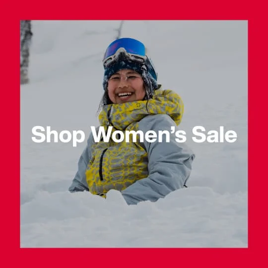 A darkened image of a Burton rider sitting in powder wearing the [ak] Kimmy Anorak in a yellow and gray print in the background. The text 'Shop Women's Sale' is in white on top.