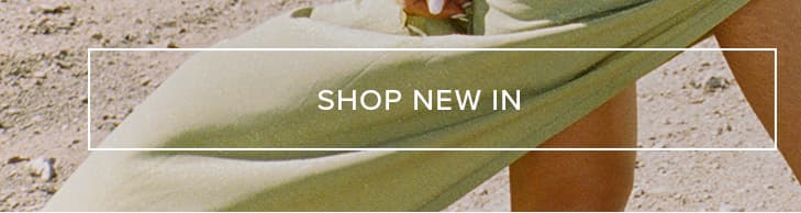 Muted sage green fabric draped over a person's leg on light brown sand. "SHOP NEW IN" to explore our latest fashion collection.