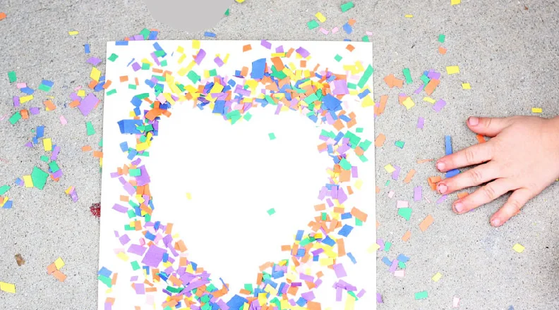 Hand sprinkling colorful confetti to create a heart-shaped design on paper.