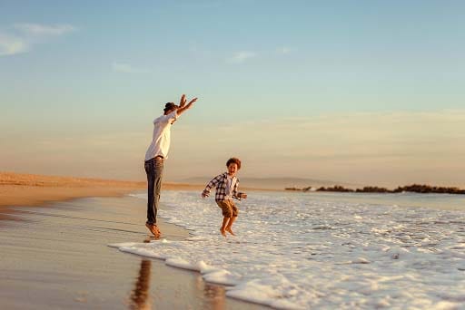 A man and a young boy joyfully play at the edge of the ocean, jumping over waves on a sandy beach during sunset, with the sky glowing in warm tones.