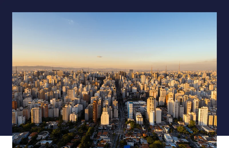 Panoramic view of a modern city skyline with numerous high-rise buildings along the waterfront, framed by lush green trees in the foreground, with the ocean visible in the background.