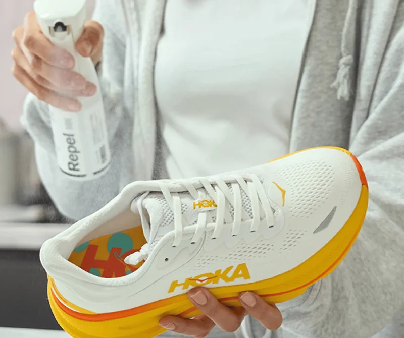 A medium shot shows a person holding a white HOKA running shoe with yellow and orange accents, while spraying it with a white bottle labeled 'Repel 10...