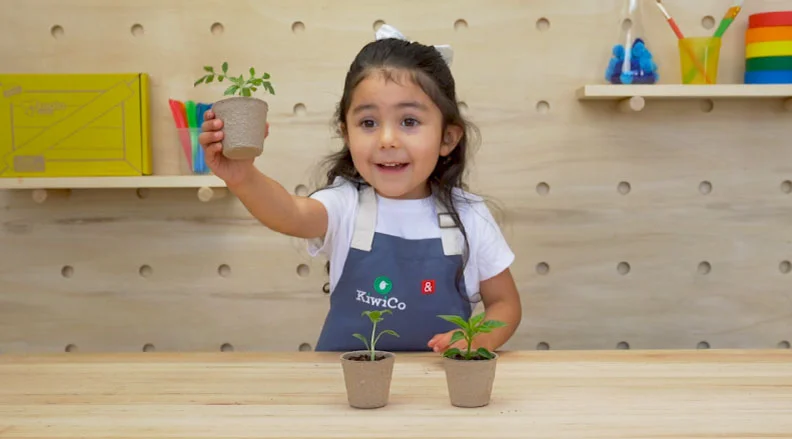 A smiling child holds a small pot with sprouting seeds, surrounded by colorful books and toys in the background. The text reads, ''Sprout Fruit Seeds – Find seeds in fruit and make them grow.''