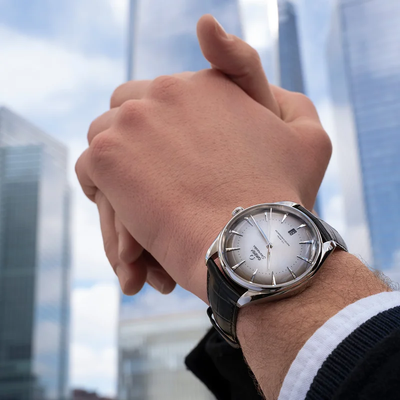 A close-up of a person’s hand wearing an OMEGA Seamaster watch with blurred background of buildings.