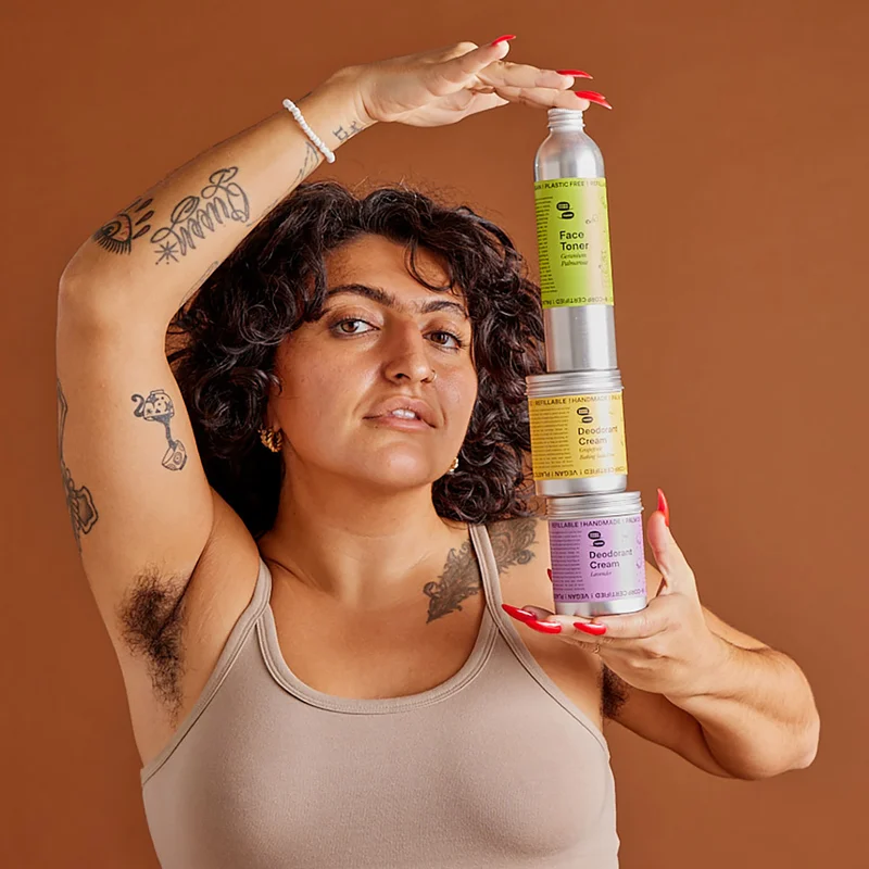 A person with dark brown curly hair and brown skin and tattoos holds a stack of bulk bottles and jars