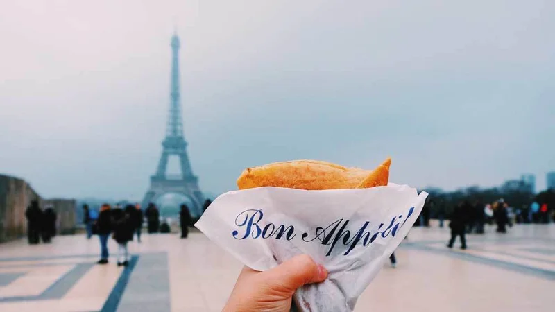 Image description: Close-up of a hand holding a crepe against the Eiffel Tower on a cloudy spring day.