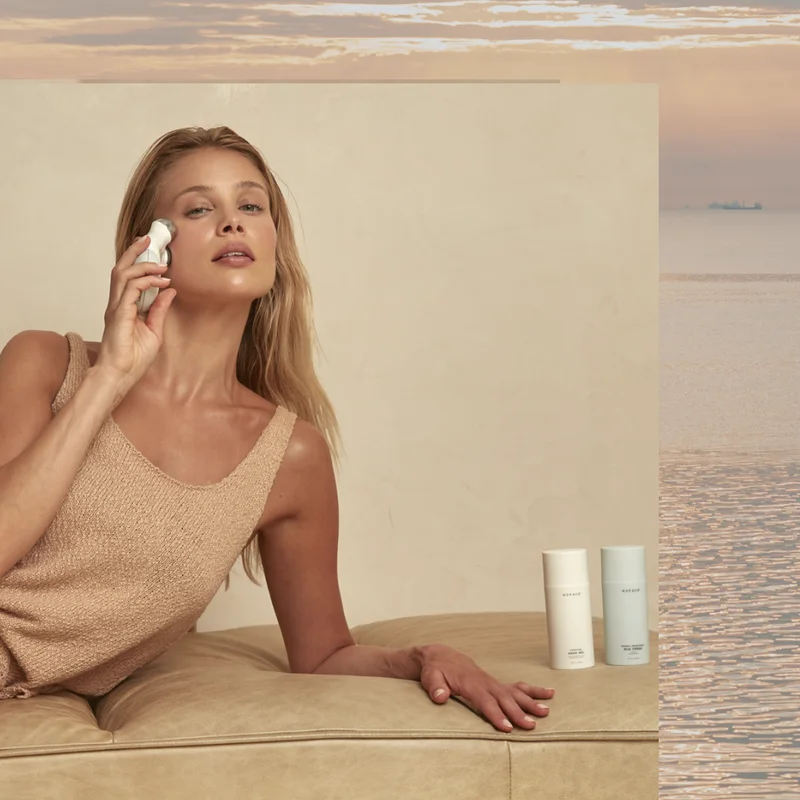 Blonde woman gliding along cheekbone with NuFACE Mini, laying down on tan chaise next to two bottles of NuFACE Microcurrent Activators, Aqua Gel and Silk Creme