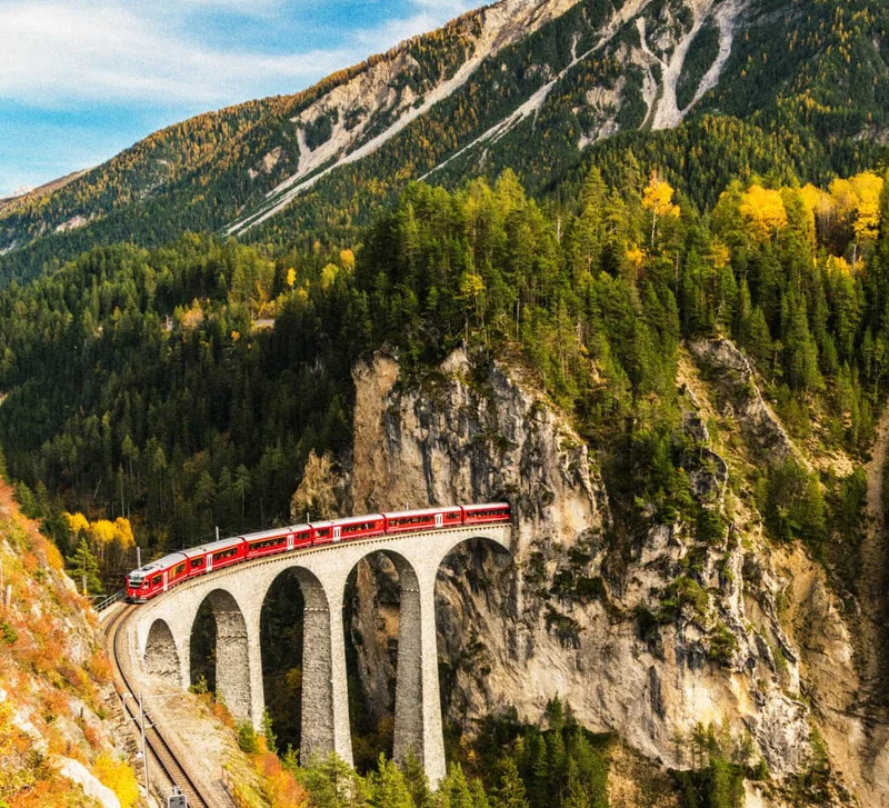 Image Description: Elevated view of the iconic Bernina Express train traveling through a lush forest in Switzerland