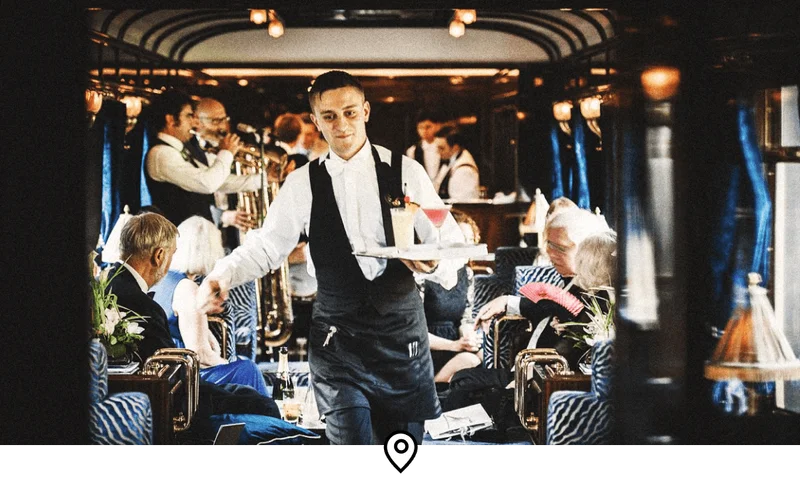 Image Description: A waiter carrying a tray of cocktails down the aisle of a luxurious train carriage, with jazz musicians playing in the background