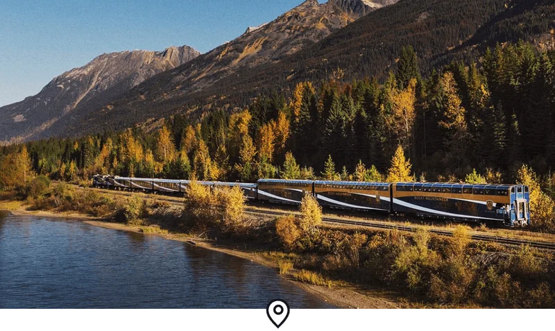 Image Description: Landscape shot of the Rocky Mountaineer train traveling between a lake and a forest-clad mountain