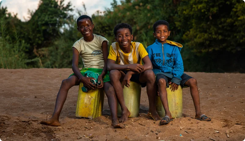 Three young boys smile at the camera sitting on top of yellow jerry cans.