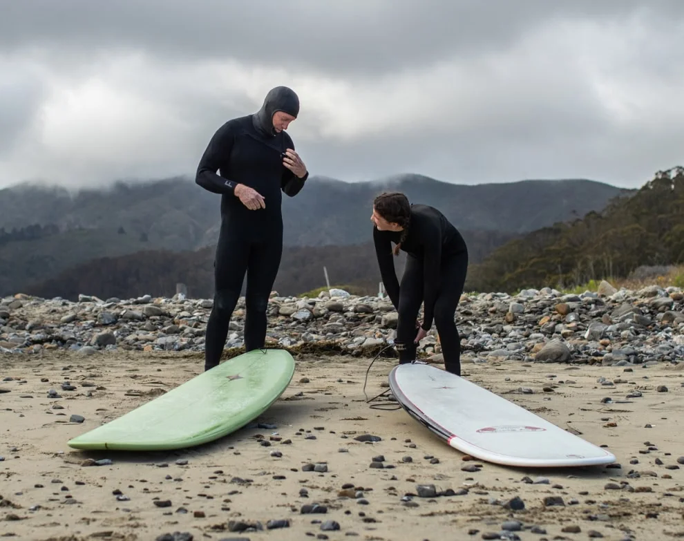 A father and daughter stand on a beach with their surfboards.