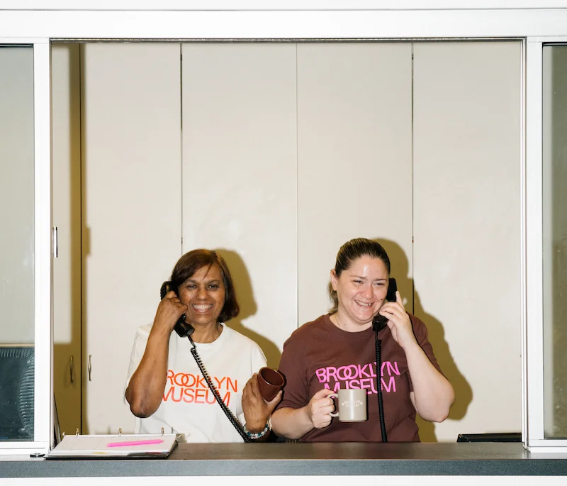 A photograph of two people wearing Brooklyn Museum t-shirts smiling and speaking on the phone