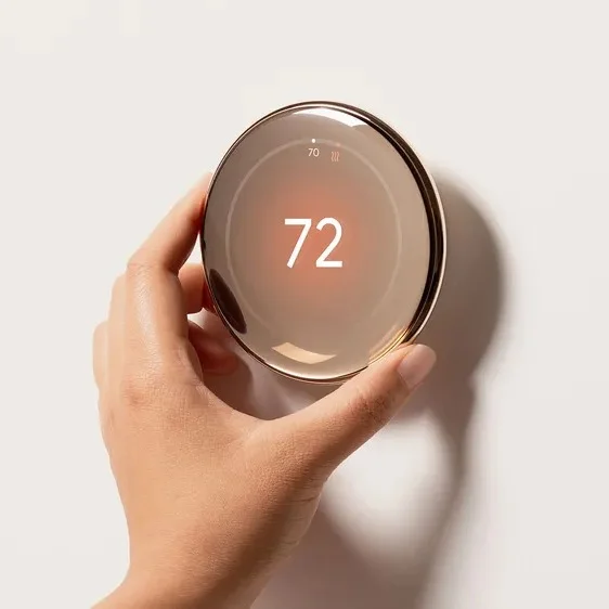 A hand reaches up from the bottom of the photo to touch a round, pebble-like Nest Thermostat. It has a shiny, domed surface and its thin border has a golden tone. The number “72” is on the screen.