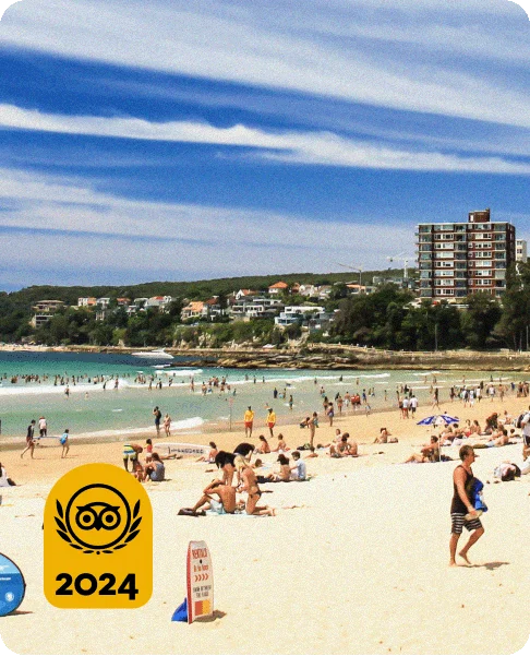 Image Description: A sandy beach with people sitting on the shore and swimming in the ocean. Green trees, a tall building, and houses are in the background. A gold Travelers’ Choice Awards Best of the Best 2024 badge is on the bottom left corner.