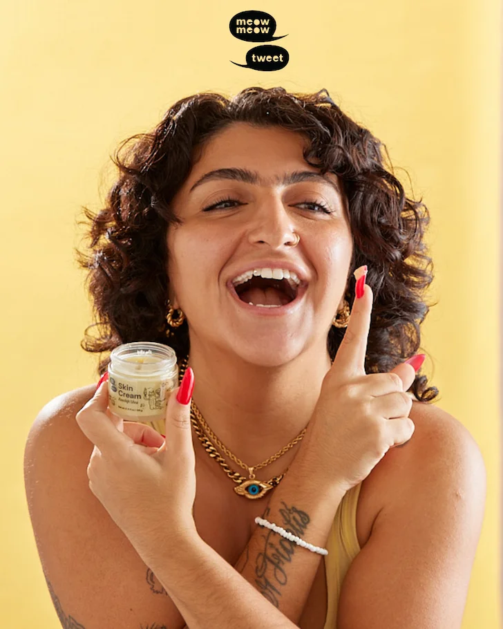 A person with tan skin and brown wavy hair smiles a huge smile at the camera as they apply skin cream to their cheek.