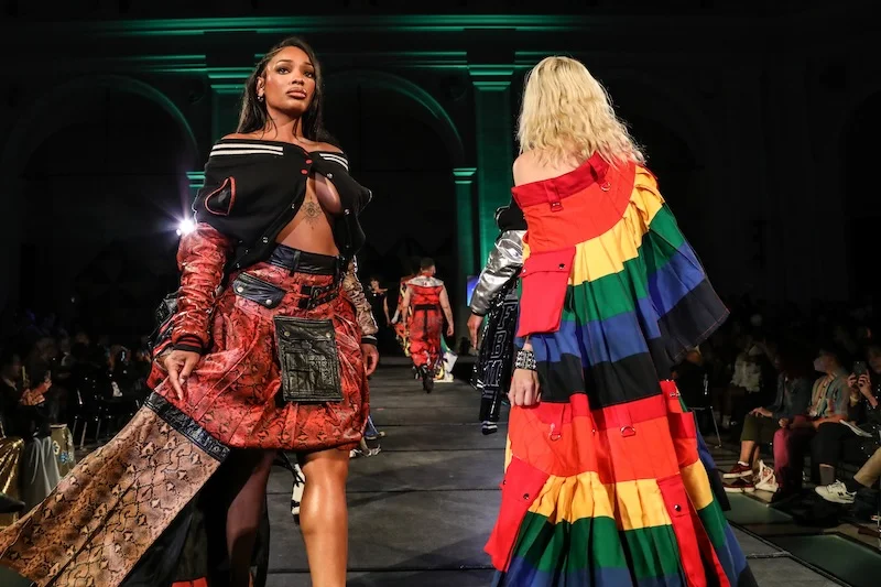 Two people on a runway, one wearing a varsity-style jacket and snake skin skirt; the other is wearing a striped rainbow dress