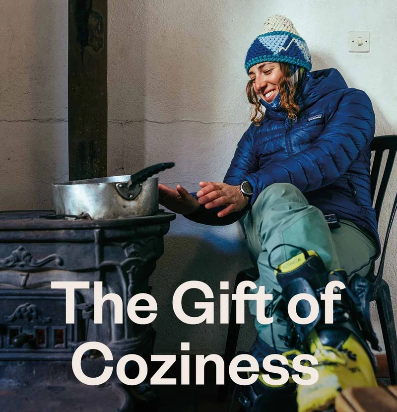 A skier warms her hands by the heat of a woodstove in a mountain hut.