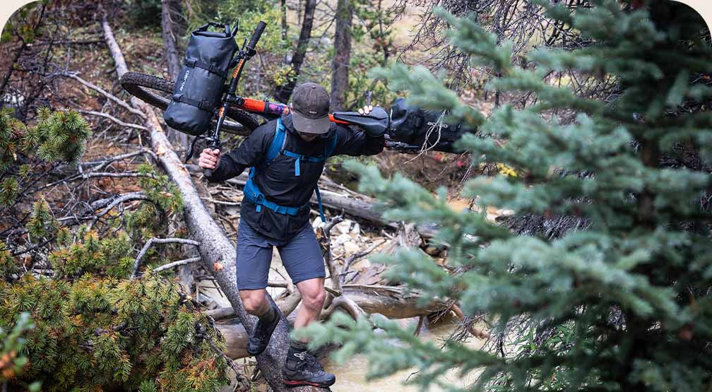 A mountain biker carries his bike over his shoulders while crossing a creek on a downed tree.