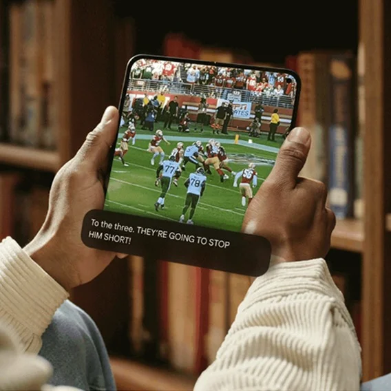 View of a person’s hands holding a Pixel Fold opened and streaming a football game. A caption box below the game reads “To the three.” Then in all caps, it reads “THEY’RE GOING TO STOP HIM SHORT!”