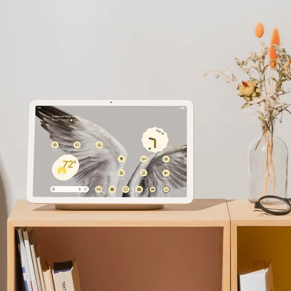 A white Google Pixel Tablet sits on its charging dock on a light brown wooden shelf. The tablet displays a colorful wallpaper and app icons.