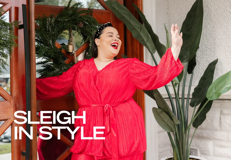 brunette woman wearing red satin shirt waving in front of door with holiday wreath