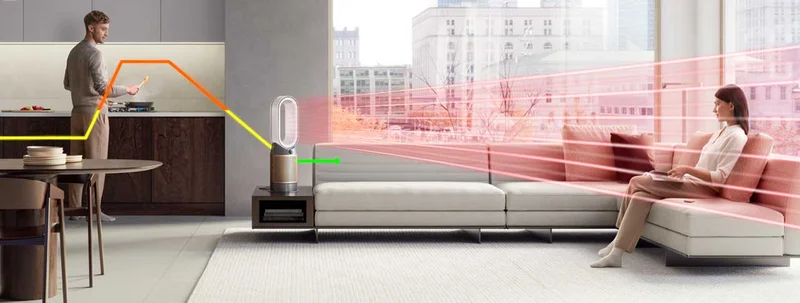 Man cooks in the kitchen with a Dyson purifier heater on Auto mode filtering pollutants from the air, shown by an air quality line. A woman nearby on the sofa is heated by the machine, shown by heating lines.
