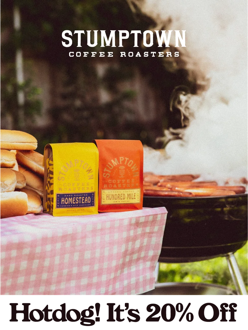Two bright Stumptown Coffee bags—Homestead and Hundred Mile—sit on a pink gingham-covered picnic table beside hot dog buns and a steaming grill, with the Stumptown Coffee Roasters logo above. Headline reads: “Hotdog! It’s 20% Off.”