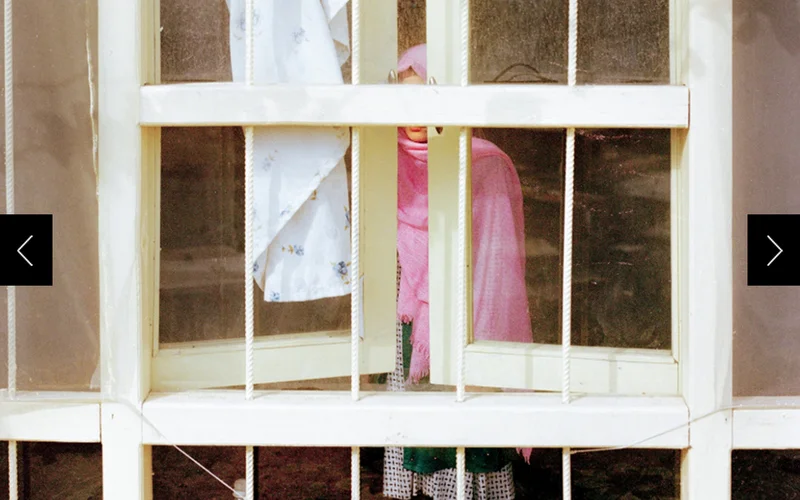 A girl in pink is photographed standing beside a window, her face obscured.