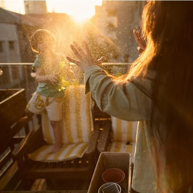 Person with long hair playing with sunlit water droplets on a balcony, with a child standing on a striped outdoor chair in the background at golden hour.