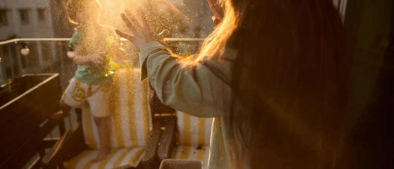 Person with long hair playing with sunlit water droplets on a balcony, with a child standing on a striped outdoor chair in the background at golden hour.