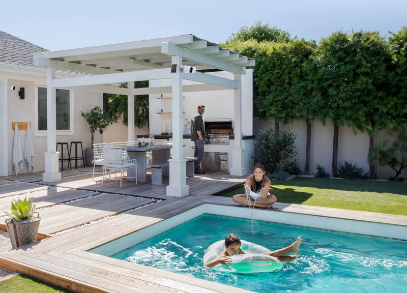 People near a pool in the back yard with various Arlo security products mounted nearby.