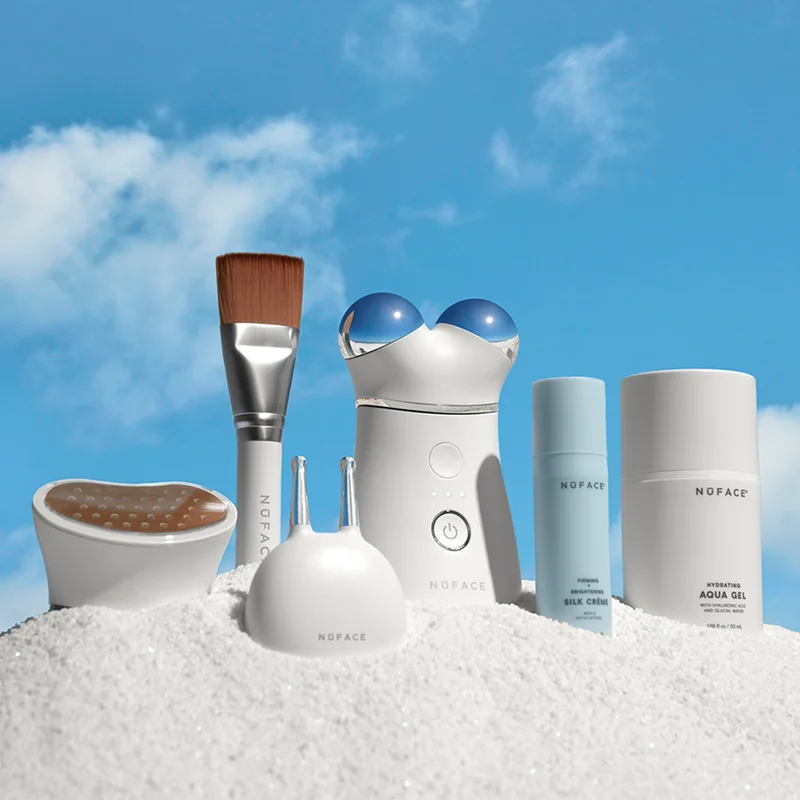 TRINITY+ Complete Set in the sand against a bright sky background. Includes Wrinkle Reducer Attachment, Effective Lip and Eye Attachment, Facial Trainer Attachment, trial-sized Silk Creme Microcurrent Activator and full-sized Aqua Gel Microcurrent Activator