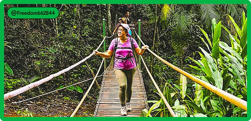 Image description: A woman wearing a backpack crosses a narrow wooden suspension bridge in a rainforest, holding onto the rails on each side.