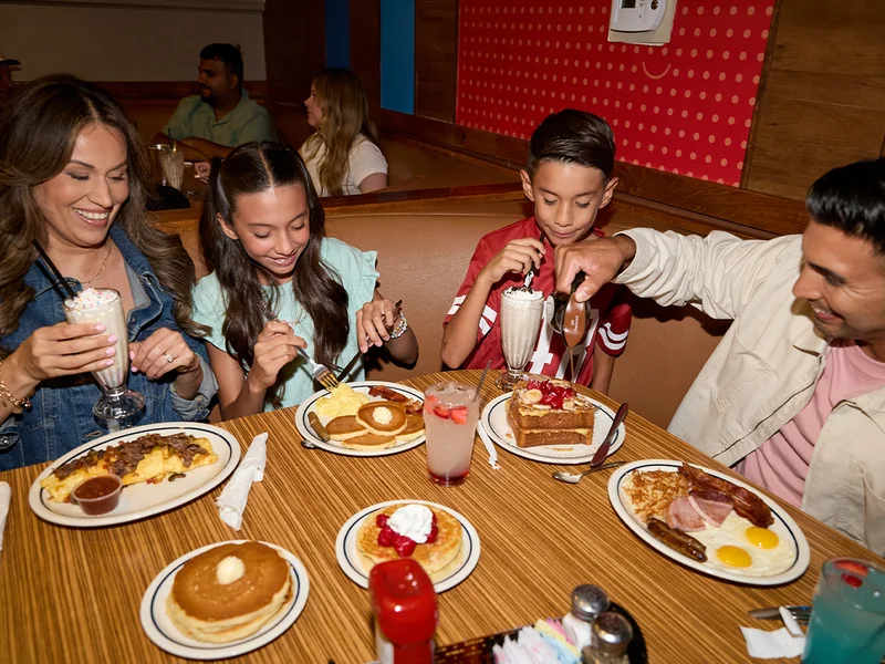 A family of four sits in a booth at IHOP with food on the table, including pancakes, French Toast and omelettes. A mom and son sip milkshakes while the daughter eats and dad pours syrup.