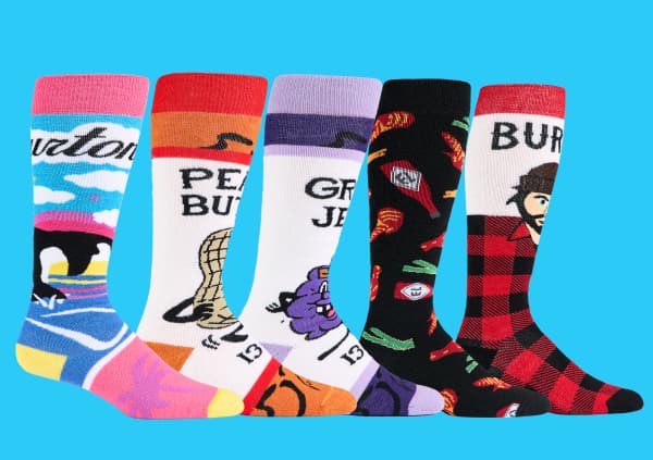 Five Burton novelty knee-high socks displayed against a light blue background, each with a different graphic print: an orca and mountain landscape, a peanut butter jar with a cobra, a grape jelly jar with a purple cartoon character, a hot chips pattern on black, and a red buffalo plaid with a bearded lumberjack portrait and Burton branding.