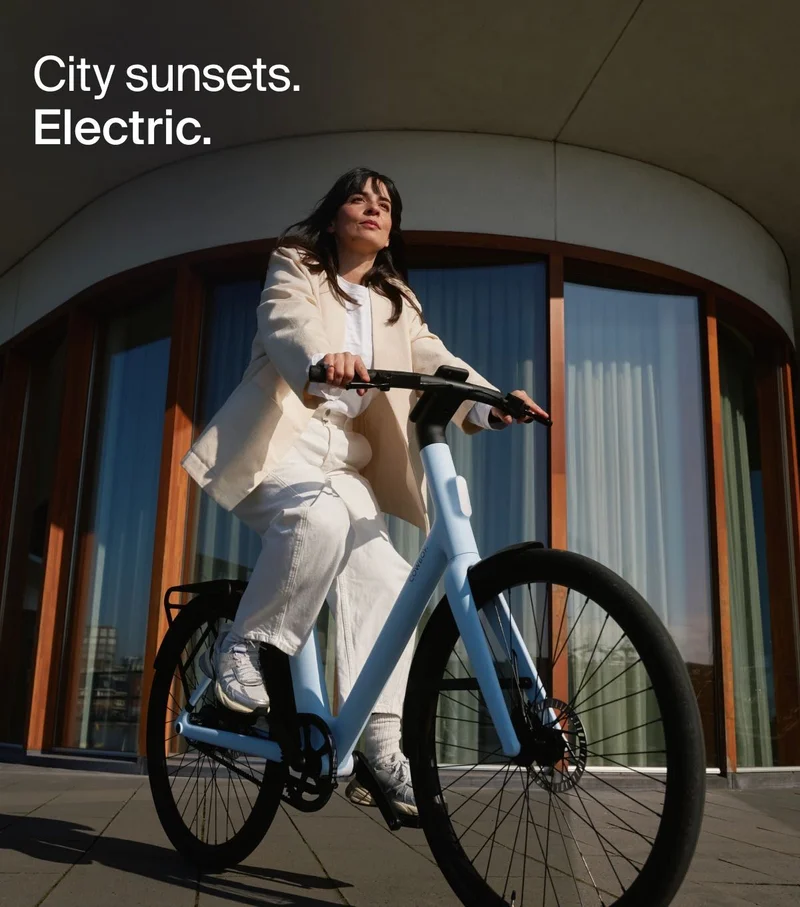 A person rides a Cowboy electric bike in the Breeze colorway, bathed in the evening light. The text overlay reads: “City Sunsets. Electric.”
