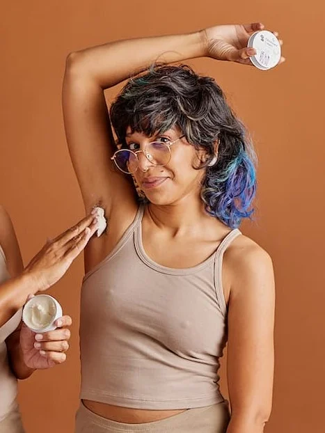 A person off screen applies deodorant cream to a person smiling with their arm up over their head.