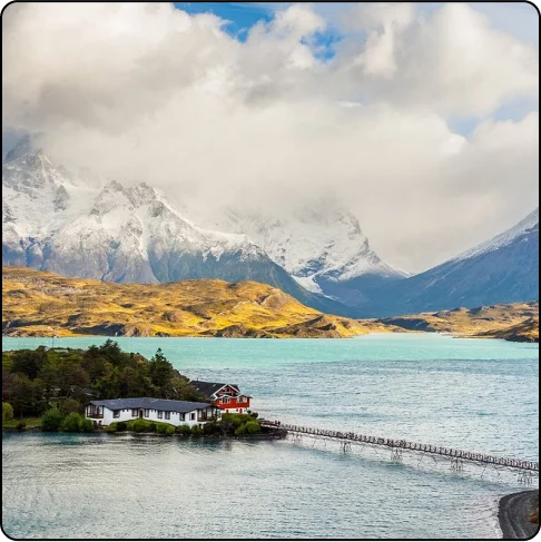Image Description: A large body of water with a small forested island and bridge in the foreground. In the background, the water turns a bright turquoise color with green rolling hills and larger snow-capped mountains in the distance.