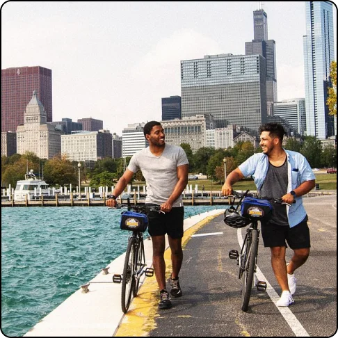 Image Description: Two people walking their bikes on a path along the water with tall city buildings in the background.