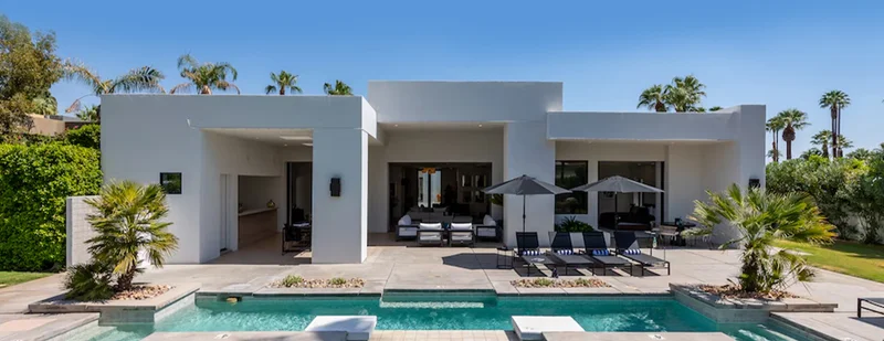 A white two-story house with a pool, sun loungers, umbrellas, palm trees, and a vibrant summer setting.