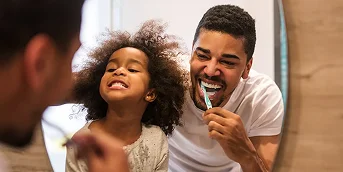 A father and child smile as they brush their teeth in the mirror.
