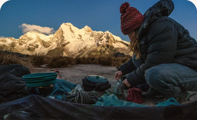 A person cooking food in camp with a large snow-covered mountain in the distance.